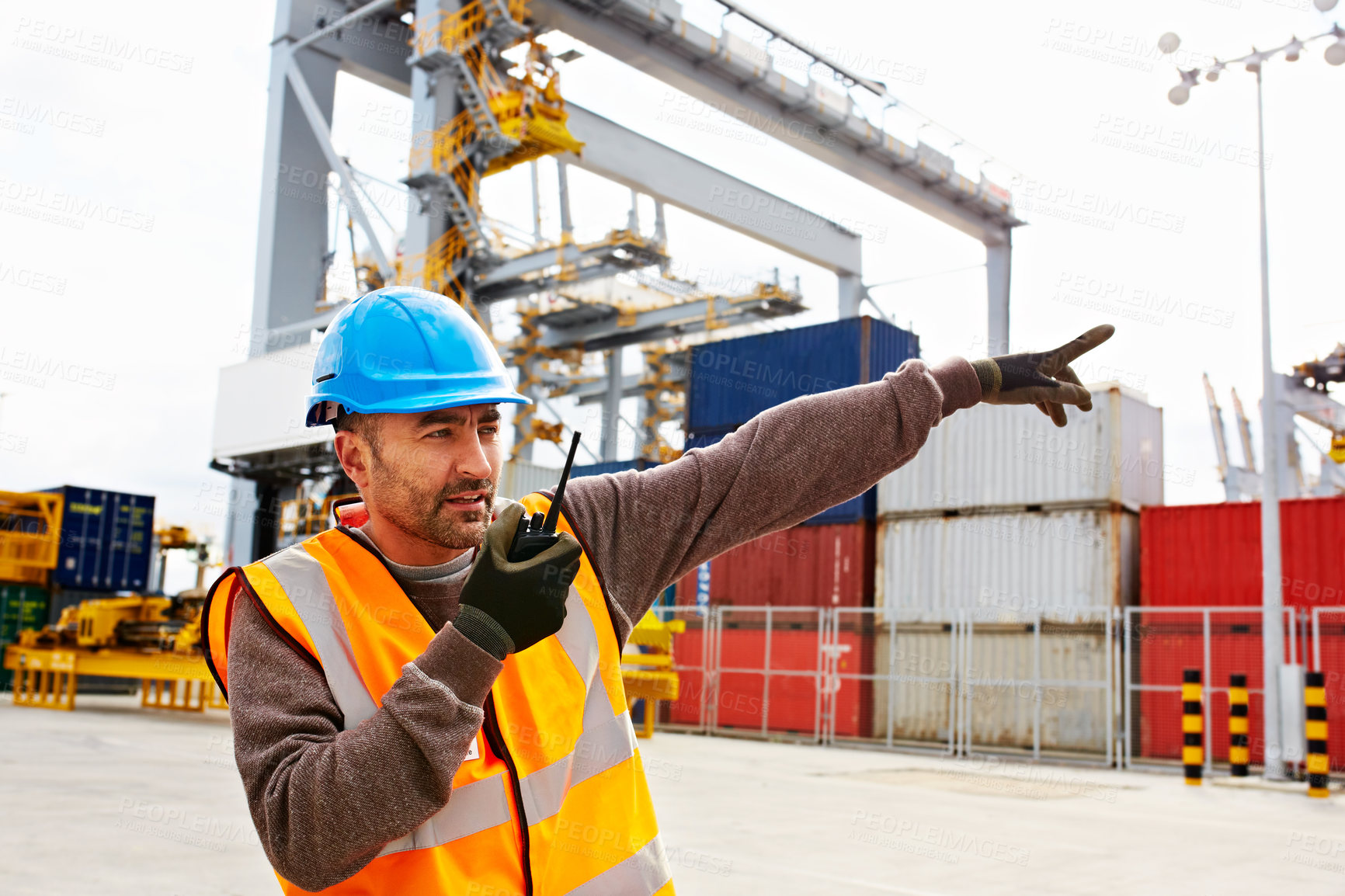 Buy stock photo Shot of a young man in workwear talking on a walkie talkie while standing on a large commercial dock