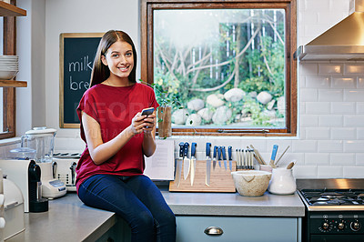 Buy stock photo Shot of a young woman sitting on her kitchen counter using a cellphone