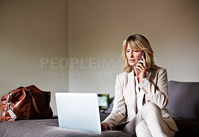 Buy stock photo Shot of a mature businesswoman sitting on a hotel bed using a laptop while talking on her cellphone