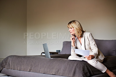 Buy stock photo Shot of a mature businesswoman sitting on a hotel bed using a laptop and reading paperwork