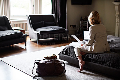 Buy stock photo Shot of a mature businesswoman sitting on a hotel bed using a laptop and reading paperwork
