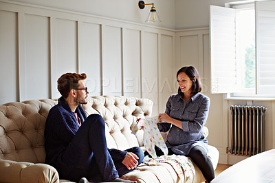 Buy stock photo Shot of a young pregnant woman showing her husband baby clothing while sitting on their sofa