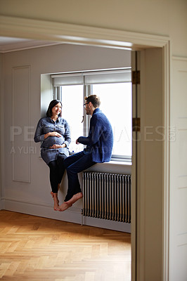 Buy stock photo Shot of a young pregnant woman talking with her husband while sitting on a window sill at home