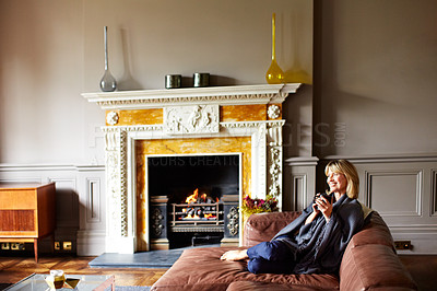 Buy stock photo Shot of a mature woman sitting on her sofa drinking tea while wrapped in a blanket
