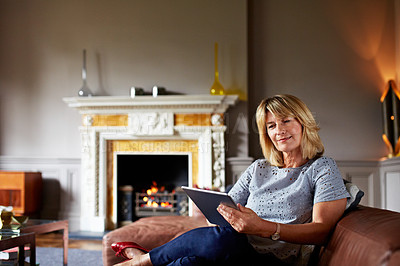 Buy stock photo Shot of a mature woman sitting on her sofa using a digital tablet with a fire glowing in the background