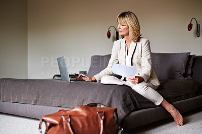 Buy stock photo Shot of a mature businesswoman sitting on a hotel bed using a laptop and reading paperwork