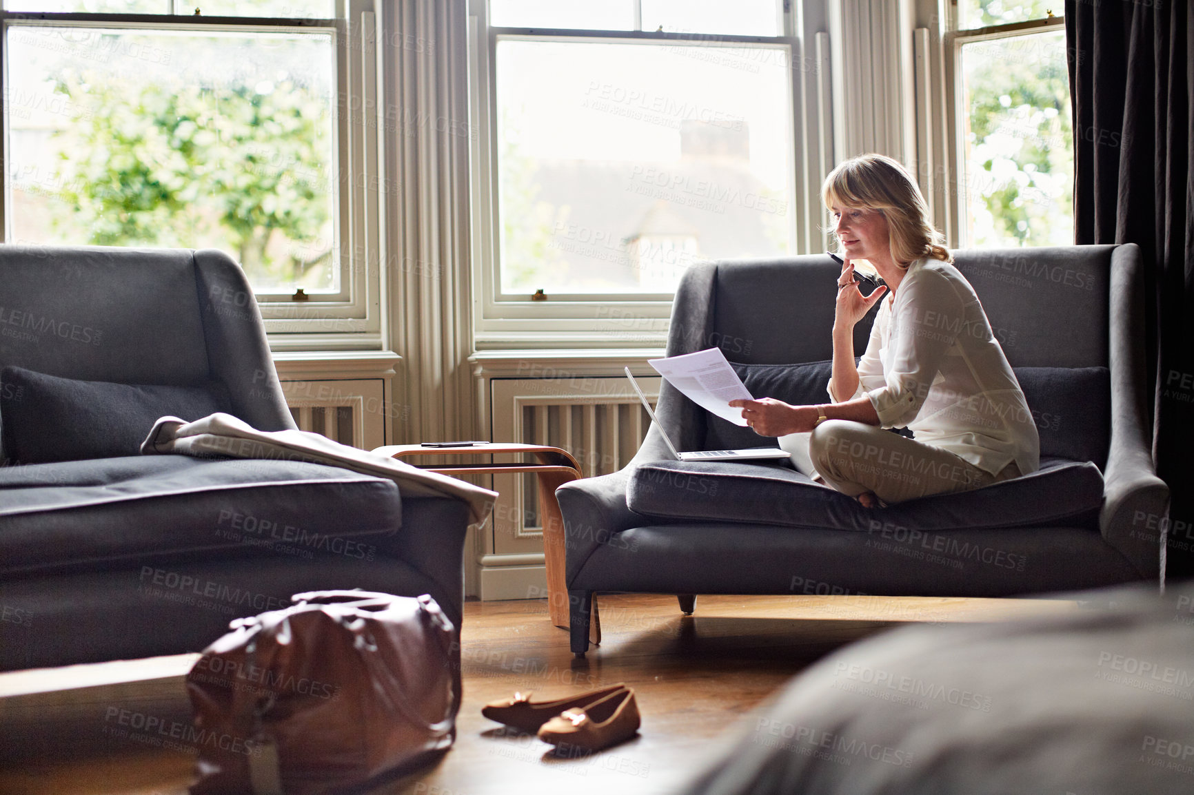Buy stock photo Shot of a mature woman working on a laptop and reading paperwork in her living room after a trip