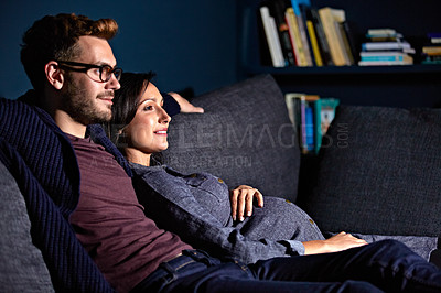 Buy stock photo Shot of a pregnant woman and her husband sitting on the sofa watching television in the evening