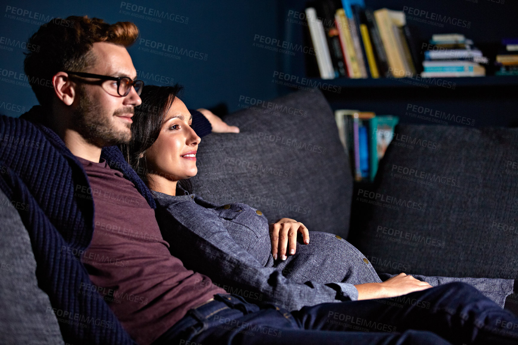 Buy stock photo Shot of a pregnant woman and her husband sitting on the sofa watching television in the evening