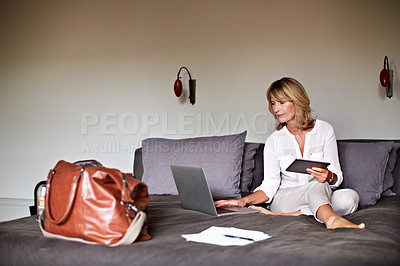 Buy stock photo Shot of a mature businesswoman sitting on a hotel bed using a laptop and digital tablet