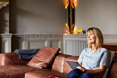 Buy stock photo Shot of a mature woman sitting on her sofa using a digital tablet with a fire glowing in the background
