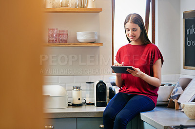 Buy stock photo Shot of a young woman sitting on her kitchen counter using a digital tablet