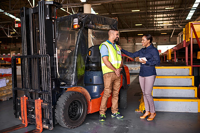 Buy stock photo Shot of two colleagues talking together beside a forklift while standing in a large warehouse