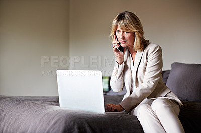Buy stock photo Shot of a mature businesswoman sitting on a hotel bed talking on her cellphone and using a laptop