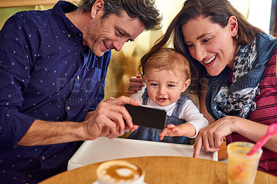 Buy stock photo Shot of a mother and father showing their baby girl photos on a phone while sitting in a cafe