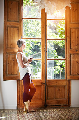 Buy stock photo Shot of a mature woman standing by a window at home drinking a glass of wine