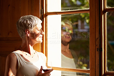 Buy stock photo Shot of a mature woman standing by a window at home drinking a glass of wine