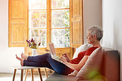 Buy stock photo Shot of a mature woman relaxing on her sofa at home using a laptop