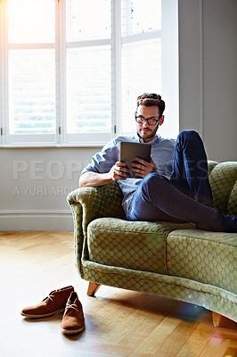 Buy stock photo Shot of a young man sitting on his sofa at home using a digital tablet