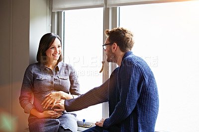 Buy stock photo Shot of a man touching his pregnant wife's stomach while sitting on a window sill at home