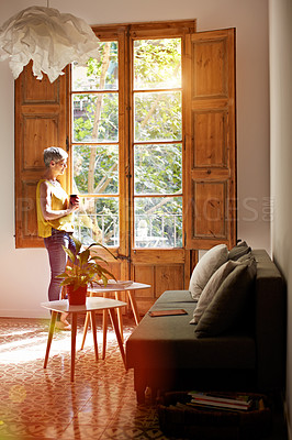 Buy stock photo Shot of a mature woman standing by a window at home drinking a cup of coffee