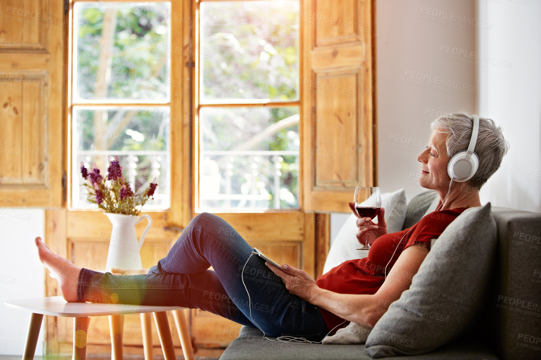 Buy stock photo Wine, headphones and mature woman on sofa with music, streaming subscription and relax on weekend. Happy, home and person with alcohol listen to radio, track and playlist for break, me time and rest