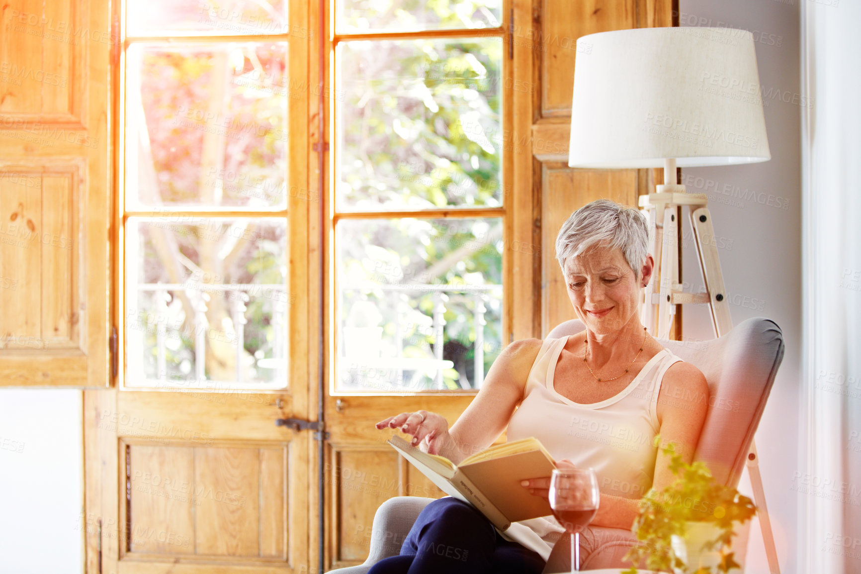 Buy stock photo Book, reading and senior woman in house with relax, calm or knowledge on novel in living room. Happy, literature and elderly person in retirement with story and wine on chair in apartment on weekend.