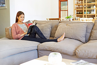 Buy stock photo Shot of a young woman sitting on her sofa reading a book and drinking a coffee