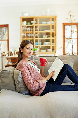 Buy stock photo Portrait of a young woman sitting on her sofa reading a book and drinking a coffee