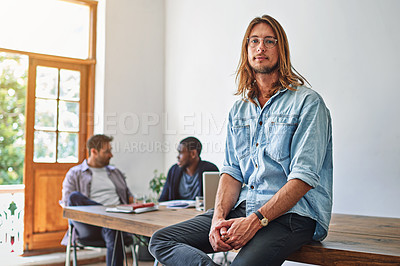 Buy stock photo Portrait of a young man sitting on a table in an office with colleagues in the background