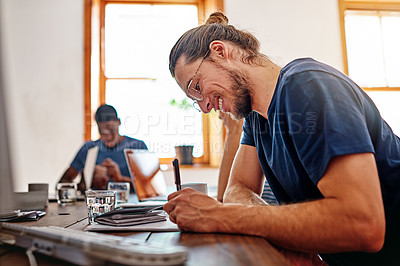 Buy stock photo Shot of a smiling young man writing in notebook while sitting at a table in an office