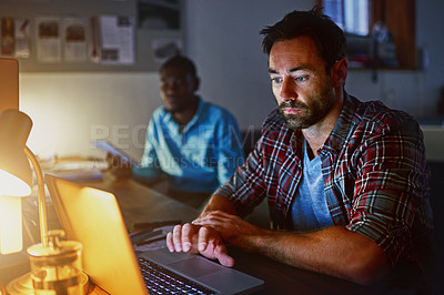 Buy stock photo Shot of a man working on a laptop in an office in the evening with colleagues in the background