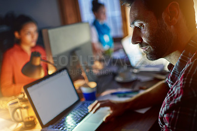 Buy stock photo Shot of a man working on a laptop in an office in the evening with colleagues in the background