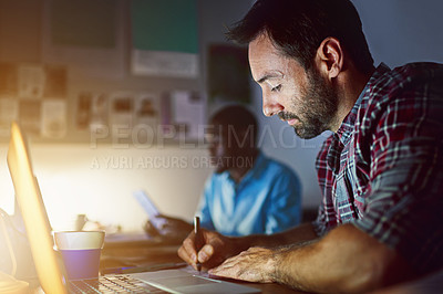 Buy stock photo Shot of a man writing notes while working on a laptop in an office late in the evening