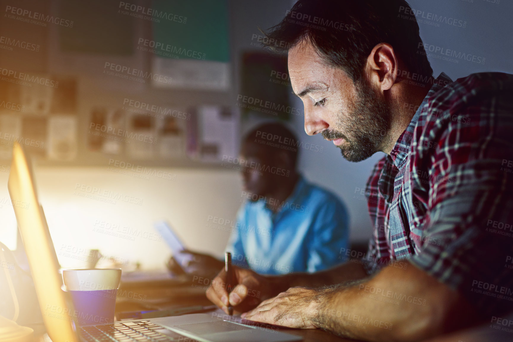 Buy stock photo Shot of a man writing notes while working on a laptop in an office late in the evening
