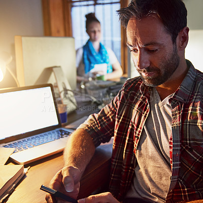Buy stock photo Shot of a man using a cellphone and working on a laptop in an office in the evening