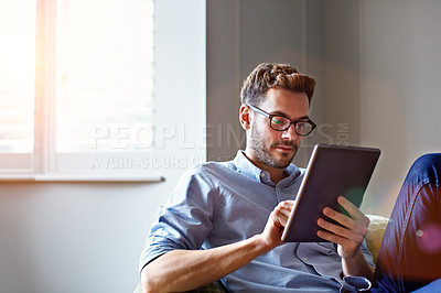 Buy stock photo Shot of a young man sitting on his sofa at home using a digital tablet