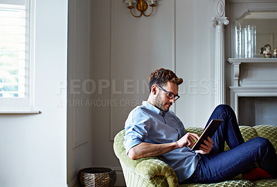 Buy stock photo Shot of a young man sitting on his sofa at home using a digital tablet