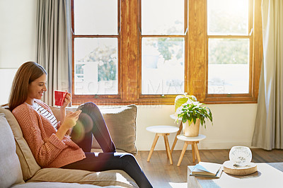 Buy stock photo Shot of a young woman sitting on her living room sofa drinking a coffee and using a cellphone