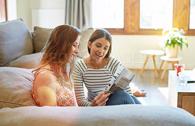 Buy stock photo Shot of two young friends sitting on the living room floor reading a magazine and talking together