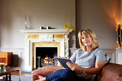 Buy stock photo Portrait of a mature woman sitting on her sofa using a digital tablet with a fire glowing in the background