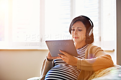 Buy stock photo Shot of a pregnant woman sitting on her sofa wearing headphones and using a digital tablet