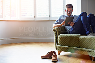 Buy stock photo Shot of a young man sitting on his sofa at home using a digital tablet