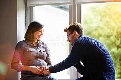 Buy stock photo Shot of a man touching his pregnant wife's stomach while sitting together on a window sill at home