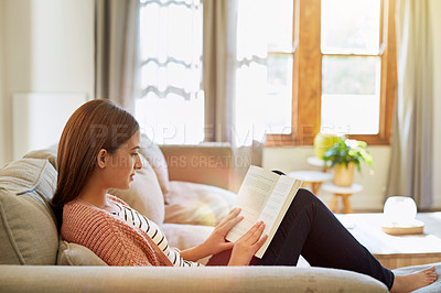 Buy stock photo Shot of a smiling young woman sitting on her sofa reading a book