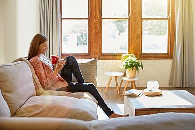 Buy stock photo Shot of a young woman sitting on her living room sofa drinking a coffee and using a cellphone