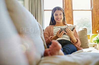 Buy stock photo Portrait of a smiling young woman sitting on her sofa watching a movie on a digital tablet with headphones