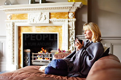 Buy stock photo Shot of a mature woman sitting on her sofa drinking tea while wrapped in a blanket