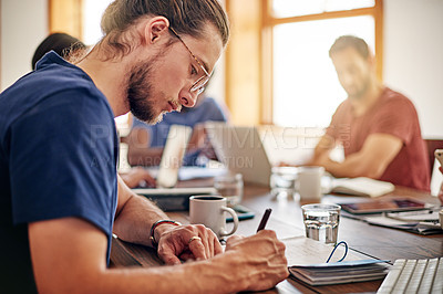 Buy stock photo Shot of a young man writing in notebook while sitting at a table in an office with colleagues in the background