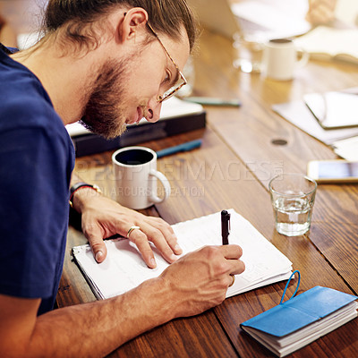 Buy stock photo Shot of a smiling young man writing in notebook while sitting at a table in an office